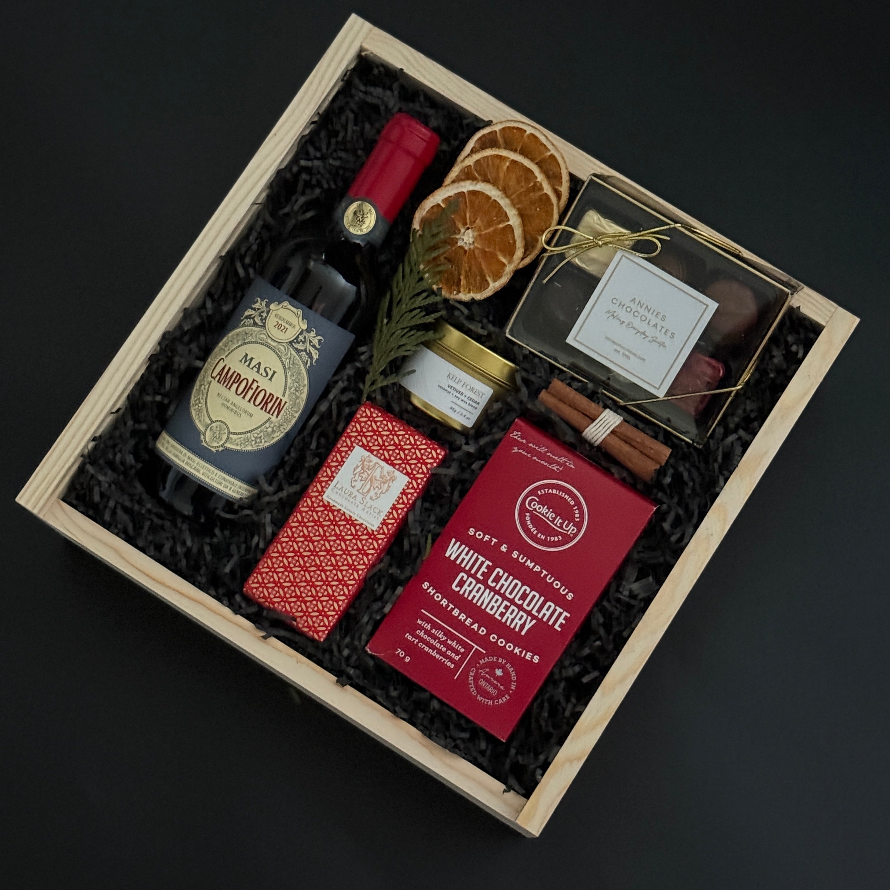 Gift box with a bottle of wine, chocolate bars, and dried oranges in a wooden box on a dark background.
