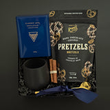 The Midnight Cocoa Gift Box displayed with artisan drinking chocolate, matte black stoneware cup, gold stirring spoon, dark chocolate pretzels, and festive cinnamon accents on a soft winter backdrop.