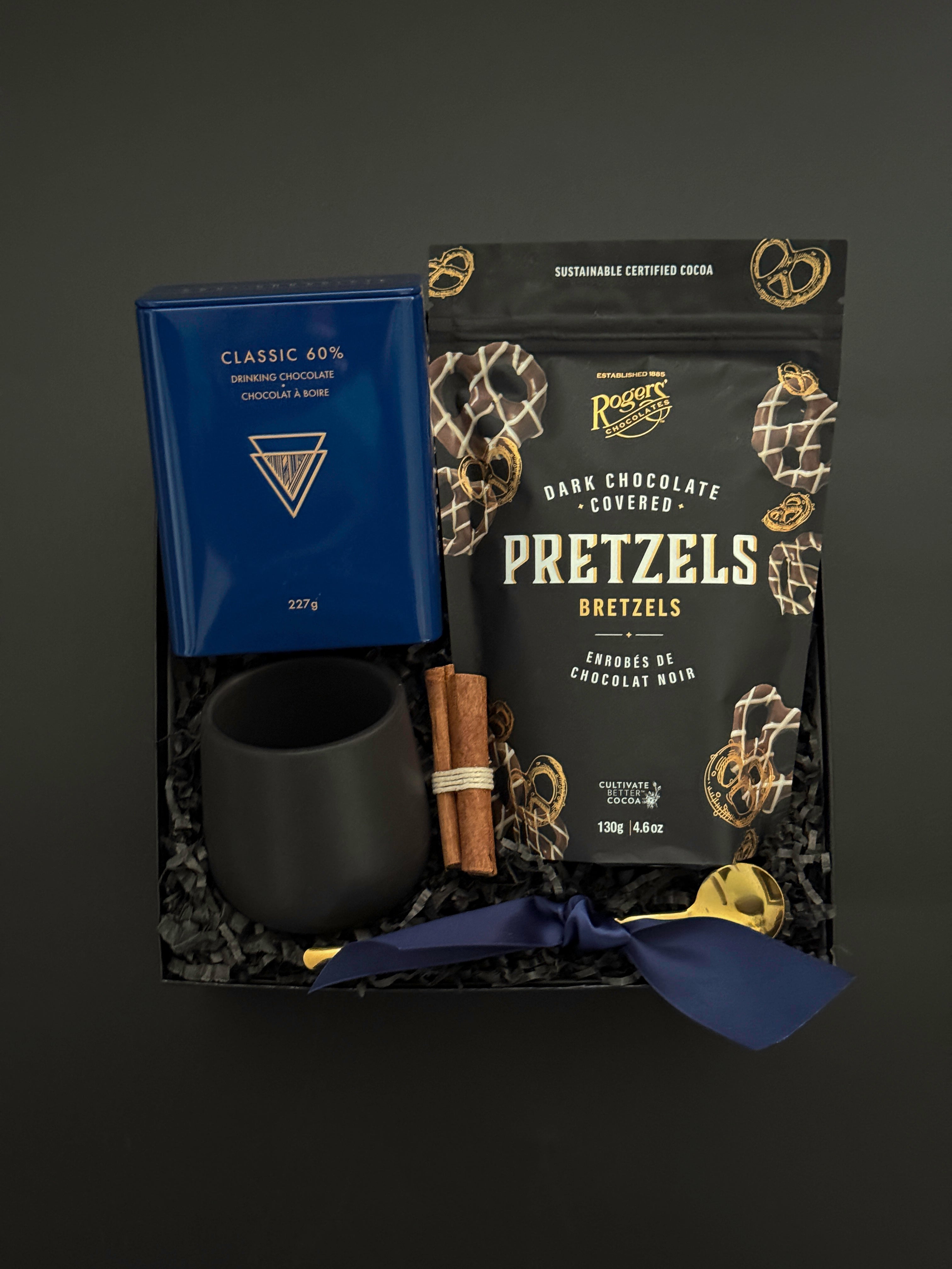 The Midnight Cocoa Gift Box displayed with artisan drinking chocolate, matte black stoneware cup, gold stirring spoon, dark chocolate pretzels, and festive cinnamon accents on a soft winter backdrop.