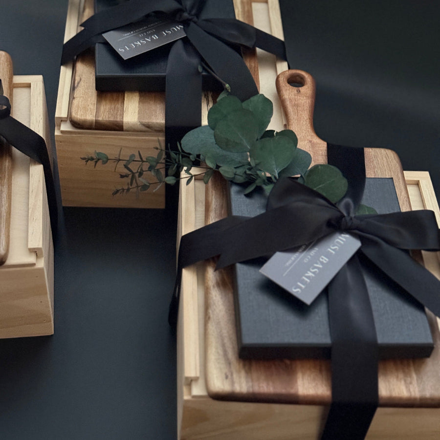 Elegant artisan gift boxes with wooden charcuterie boards, black ribbon, eucalyptus greenery, and premium packaging from Muse Baskets