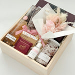 Gift box with bath products, a small bouquet, and boxed chocolates in wood basket and on a white background