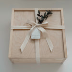 A square light wood presentation box tied with a cream ribbon and bow, with a small eucalyptus sprig, photographed from above on a light neutral background.