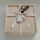 A square light wood presentation box tied with a cream ribbon and bow, with a small eucalyptus sprig, photographed from above on a light neutral background.