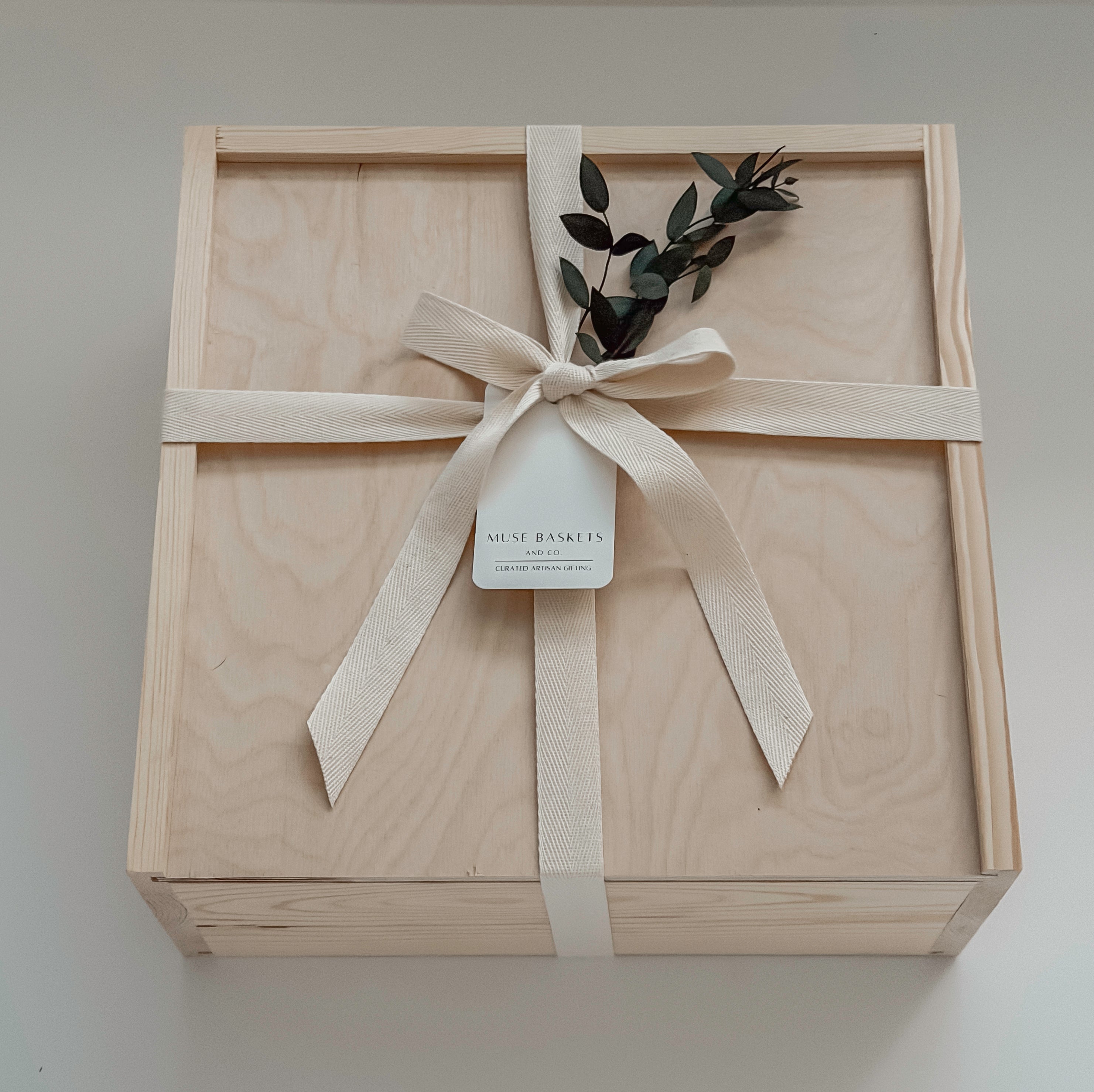 A square light wood presentation box tied with a cream ribbon and bow, with a small eucalyptus sprig, photographed from above on a light neutral background.