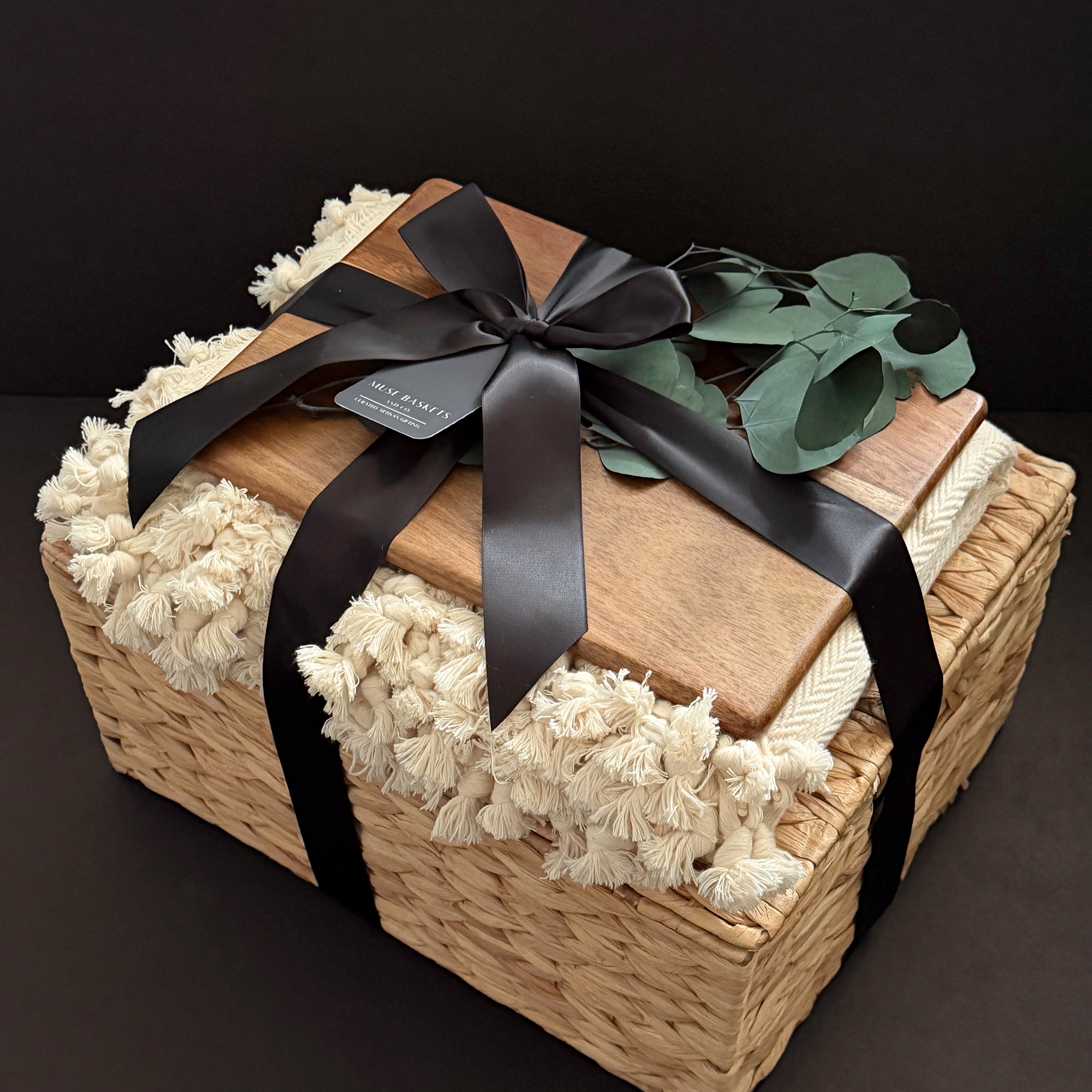 A woven wicker gift basket with a wooden lid, wrapped in a black ribbon and bow, and lined with cream-colored fringed fabric. A small tag on the ribbon reads 'The Gift Shop'.