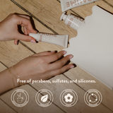 Hand holding a tube of cream on a wooden floor with product packaging in the background, featuring text about product ingredients and certification logos.