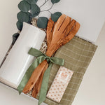 Decorative set with a white candle, wooden spoon, green ribbon, and a checkered cloth on a white surface.