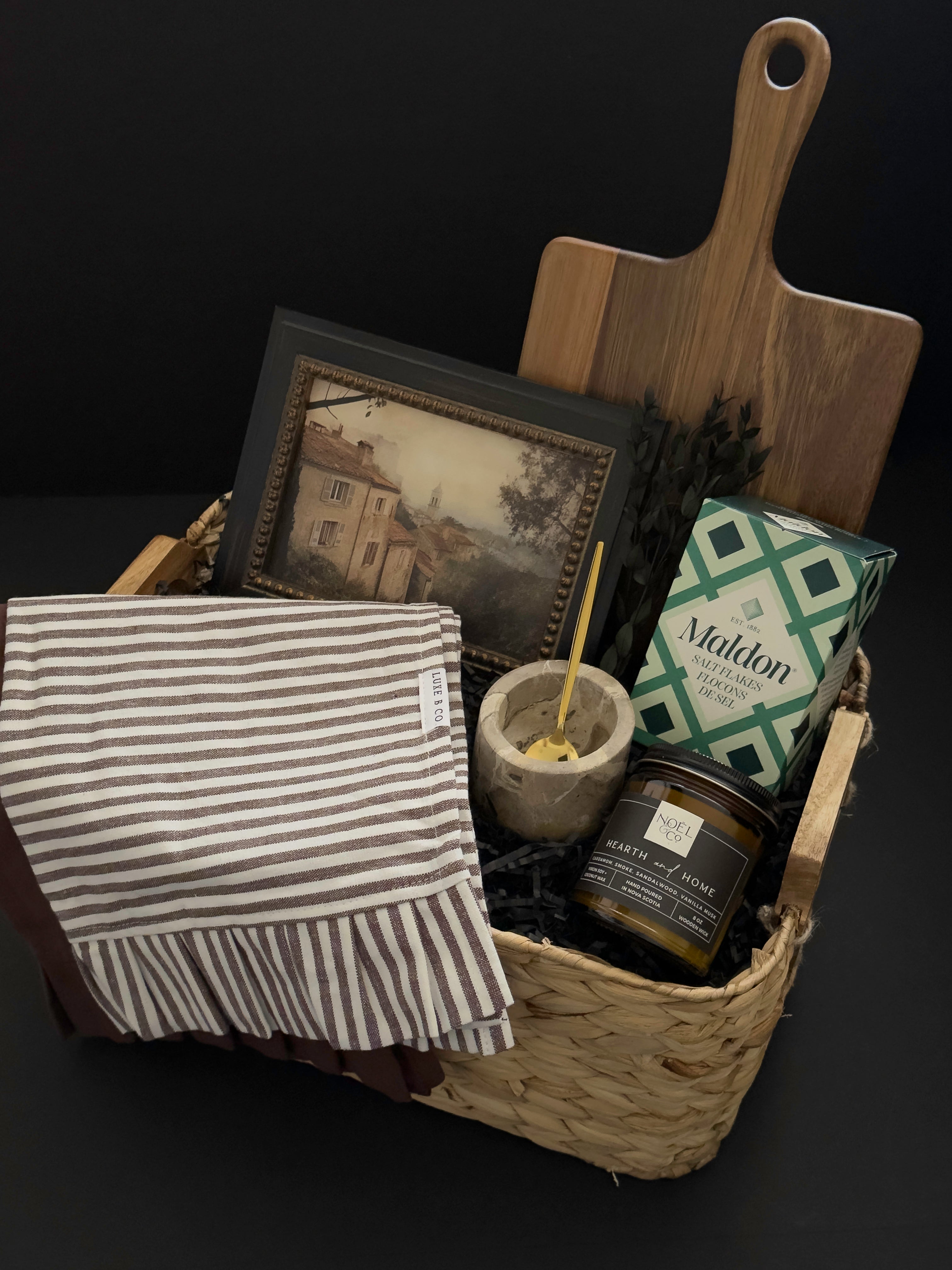 Realtor possession day gift featuring framed vintage-style artwork, wood serving board, striped kitchen towel, candle and pantry accents in a woven keepsake basket.