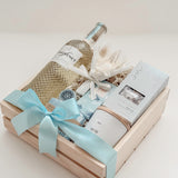 Seraphine bridal gift box by Muse Baskets and Co. in a wood crate with a satin baby blue bow, featuring Freixenet Pinot Grigio, Sealuxe Ocean Goddess bath tea, a Muse candle by Aija, olive and bloom soap, lip balm, and a white preserved floral bouquet, a refined wedding, engagement, and bridal shower gift.