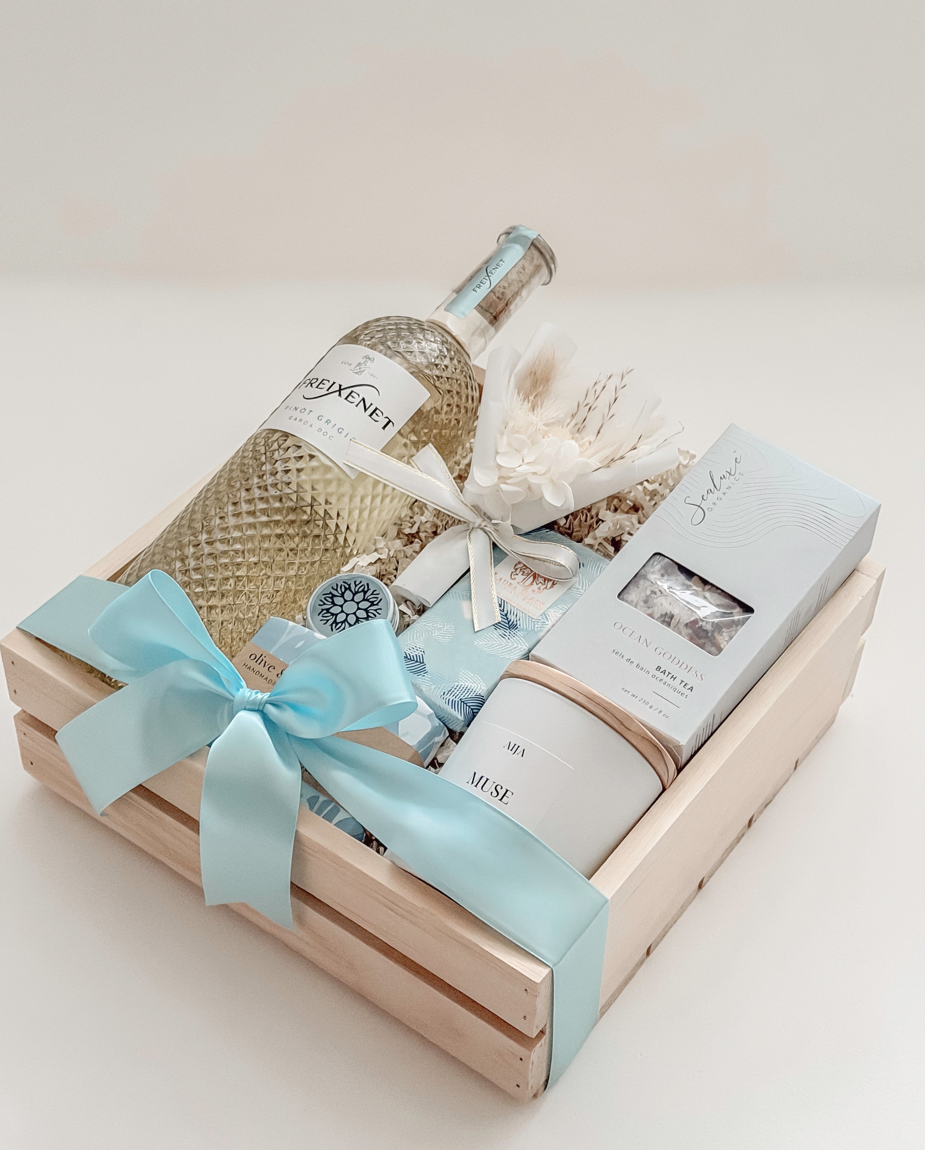 Seraphine bridal gift box by Muse Baskets and Co. in a wood crate with a satin baby blue bow, featuring Freixenet Pinot Grigio, Sealuxe Ocean Goddess bath tea, a Muse candle by Aija, olive and bloom soap, lip balm, and a white preserved floral bouquet, a refined wedding, engagement, and bridal shower gift.
