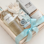Seraphine bridal gift box by Muse Baskets and Co. in a wood crate with a satin baby blue bow, featuring Freixenet Pinot Grigio, Sealuxe Ocean Goddess bath tea, a Muse candle by Aija, olive and bloom soap, lip balm, and a white preserved floral bouquet, a refined wedding, engagement, and bridal shower gift.