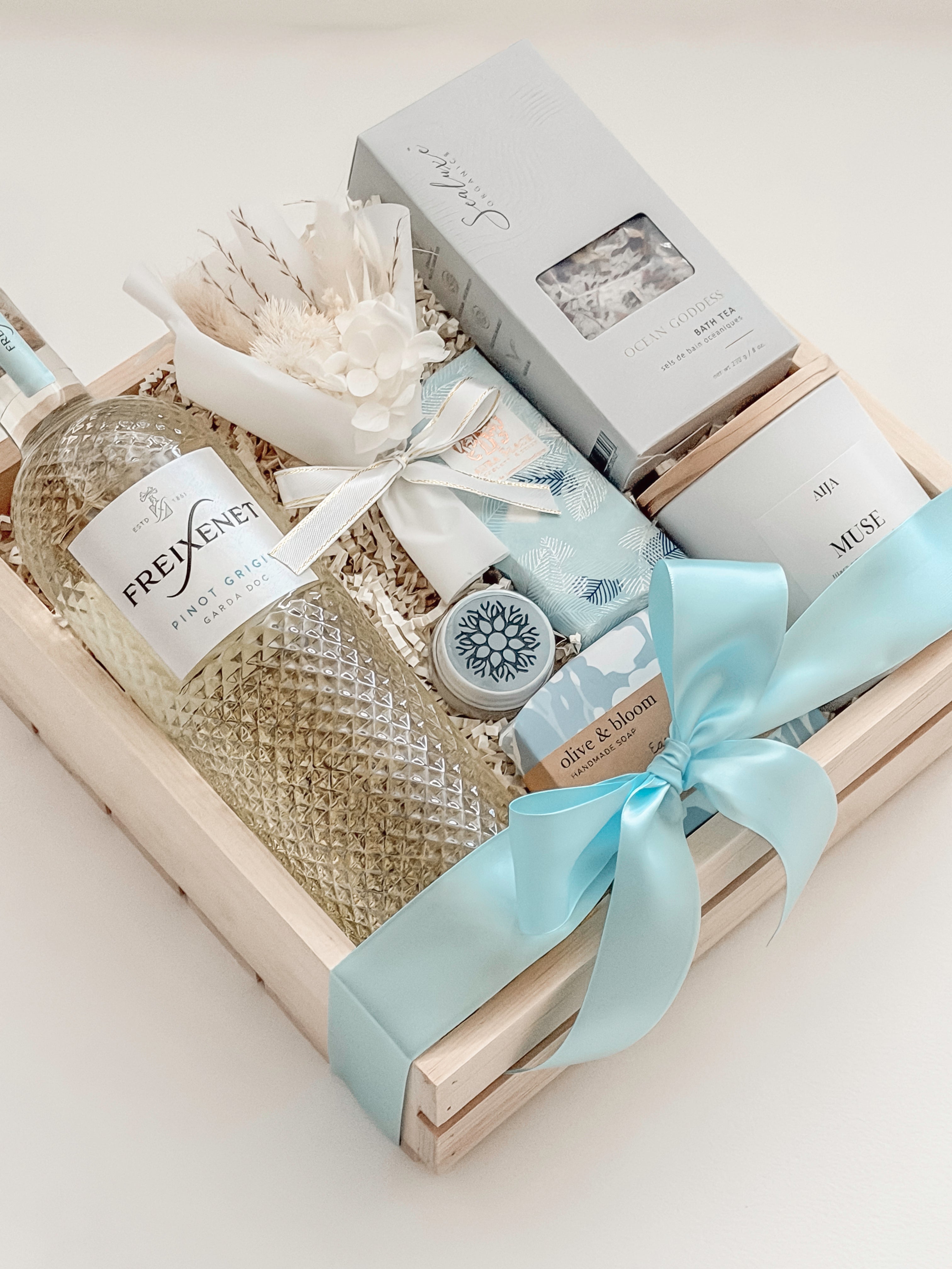 Seraphine bridal gift box by Muse Baskets and Co. in a wood crate with a satin baby blue bow, featuring Freixenet Pinot Grigio, Sealuxe Ocean Goddess bath tea, a Muse candle by Aija, olive and bloom soap, lip balm, and a white preserved floral bouquet, a refined wedding, engagement, and bridal shower gift.