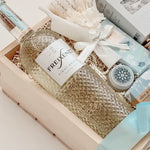 Seraphine bridal gift box by Muse Baskets and Co. shown from above, with Freixenet Pinot Grigio, Sealuxe Ocean Goddess bath tea, a Muse candle, olive and bloom handmade soap, artisan chocolate, lip balm, and a white preserved floral bouquet in a wood crate tied with baby blue ribbon, a refined wedding, engagement, and bridal shower gift from Canadian makers.