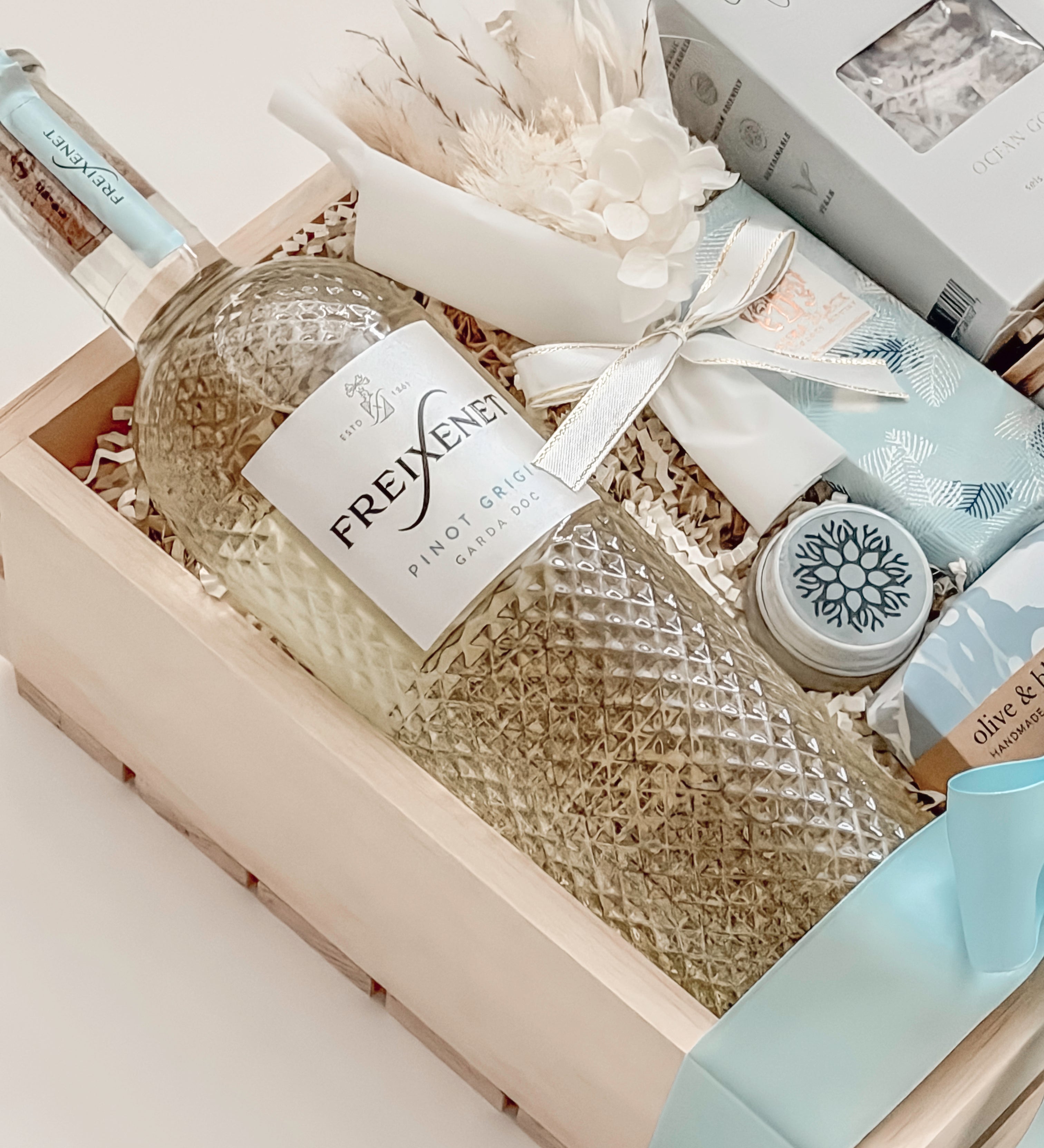 Seraphine bridal gift box by Muse Baskets and Co. shown from above, with Freixenet Pinot Grigio, Sealuxe Ocean Goddess bath tea, a Muse candle, olive and bloom handmade soap, artisan chocolate, lip balm, and a white preserved floral bouquet in a wood crate tied with baby blue ribbon, a refined wedding, engagement, and bridal shower gift from Canadian makers.