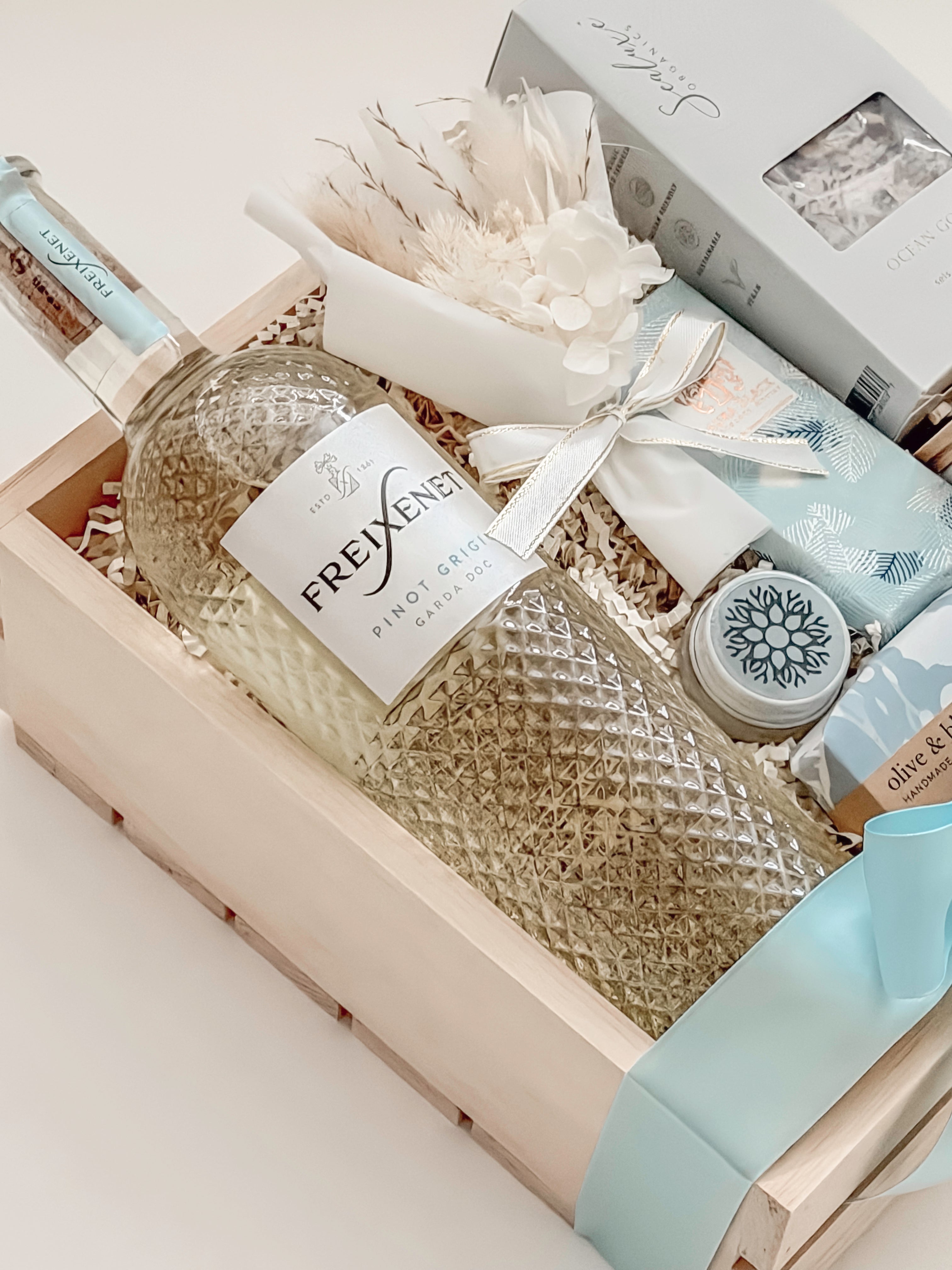 Seraphine bridal gift box by Muse Baskets and Co. shown from above, with Freixenet Pinot Grigio, Sealuxe Ocean Goddess bath tea, a Muse candle, olive and bloom handmade soap, artisan chocolate, lip balm, and a white preserved floral bouquet in a wood crate tied with baby blue ribbon, a refined wedding, engagement, and bridal shower gift from Canadian makers.