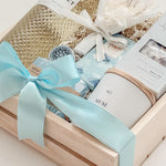 Seraphine bridal gift box by Muse Baskets and Co. in a wood crate with a satin baby blue bow, featuring Freixenet Pinot Grigio, Sealuxe Ocean Goddess bath tea, a Muse candle by Aija, olive and bloom soap, lip balm, and a white preserved floral bouquet, a refined wedding, engagement, and bridal shower gift.