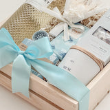 Seraphine bridal gift box by Muse Baskets and Co. in a wood crate with a satin baby blue bow, featuring Freixenet Pinot Grigio, Sealuxe Ocean Goddess bath tea, a Muse candle by Aija, olive and bloom soap, lip balm, and a white preserved floral bouquet, a refined wedding, engagement, and bridal shower gift.