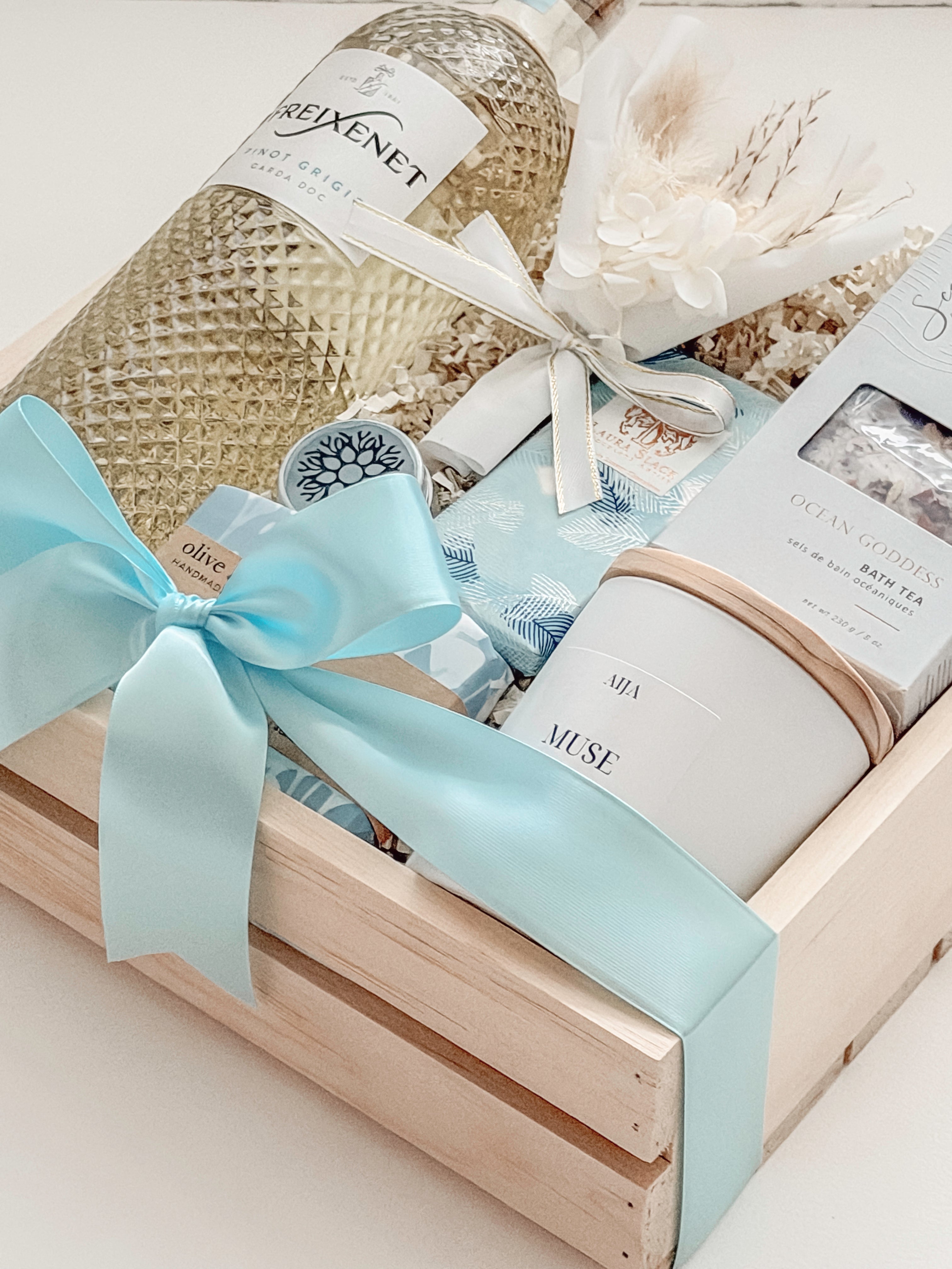 Seraphine bridal gift box by Muse Baskets and Co. in a wood crate with a satin baby blue bow, featuring Freixenet Pinot Grigio, Sealuxe Ocean Goddess bath tea, a Muse candle by Aija, olive and bloom soap, lip balm, and a white preserved floral bouquet, a refined wedding, engagement, and bridal shower gift.