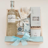 Seraphine bridal gift box by Muse Baskets and Co. in a wood crate with a satin baby blue bow, featuring Freixenet Pinot Grigio, Sealuxe Ocean Goddess bath tea, a Muse candle by Aija, olive and bloom soap, lip balm, and a white preserved floral bouquet, a refined wedding, engagement, and bridal shower gift.