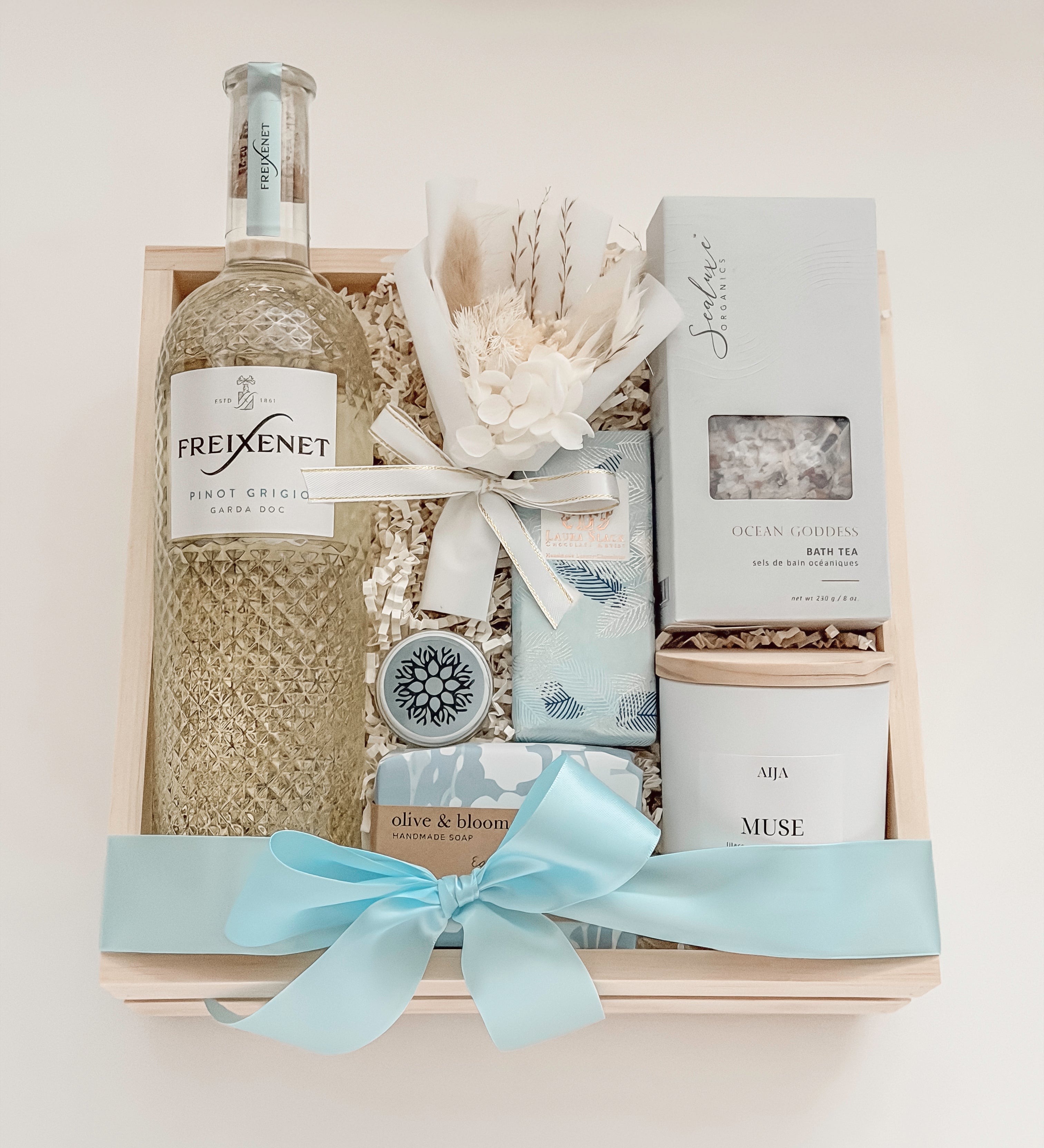 Seraphine bridal gift box by Muse Baskets and Co. in a wood crate with a satin baby blue bow, featuring Freixenet Pinot Grigio, Sealuxe Ocean Goddess bath tea, a Muse candle by Aija, olive and bloom soap, lip balm, and a white preserved floral bouquet, a refined wedding, engagement, and bridal shower gift.