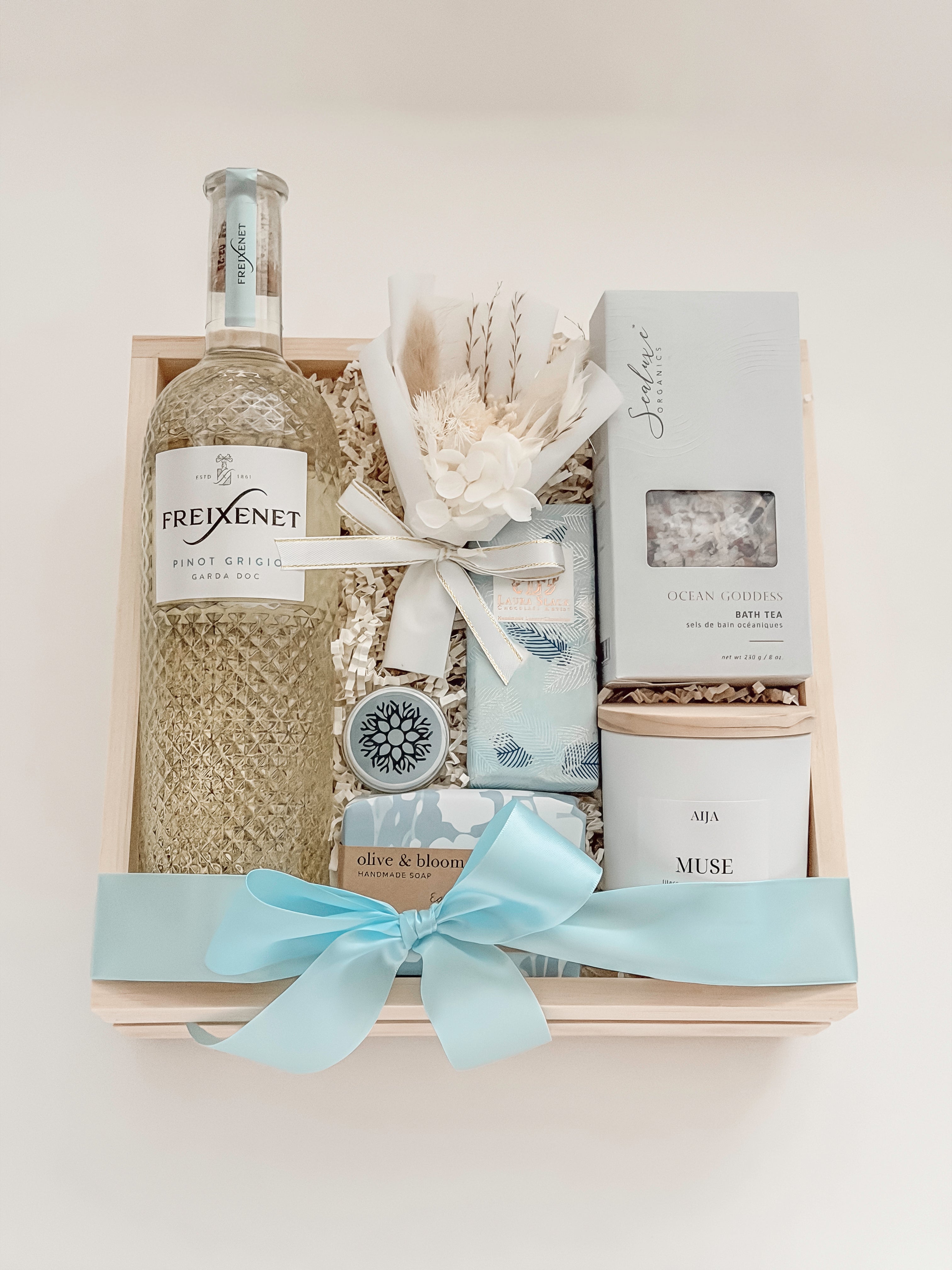 Seraphine bridal gift box by Muse Baskets and Co. in a wood crate with a satin baby blue bow, featuring Freixenet Pinot Grigio, Sealuxe Ocean Goddess bath tea, a Muse candle by Aija, olive and bloom soap, lip balm, and a white preserved floral bouquet, a refined wedding, engagement, and bridal shower gift.