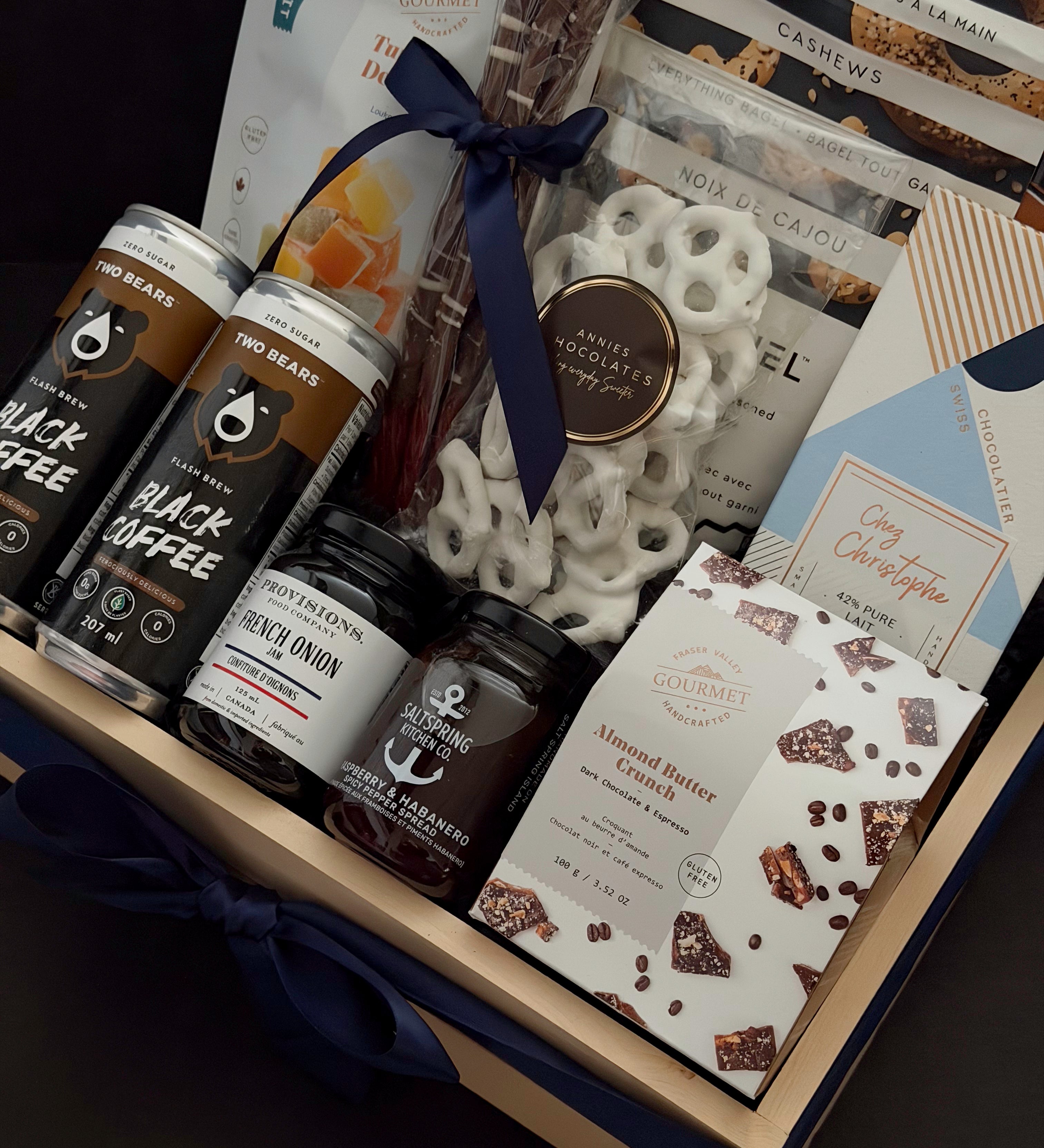 Canadian Gourmet gift box by Muse Baskets and Co., close-up of Two Bears flash brew coffee, Provisions French onion jam, Saltspring Kitchen Co. raspberry habanero spread, Fraser Valley Gourmet almond butter crunch, and white chocolate pretzels in a navy-ribboned wood crate, a luxury Canadian gift for closings and corporate occasions.