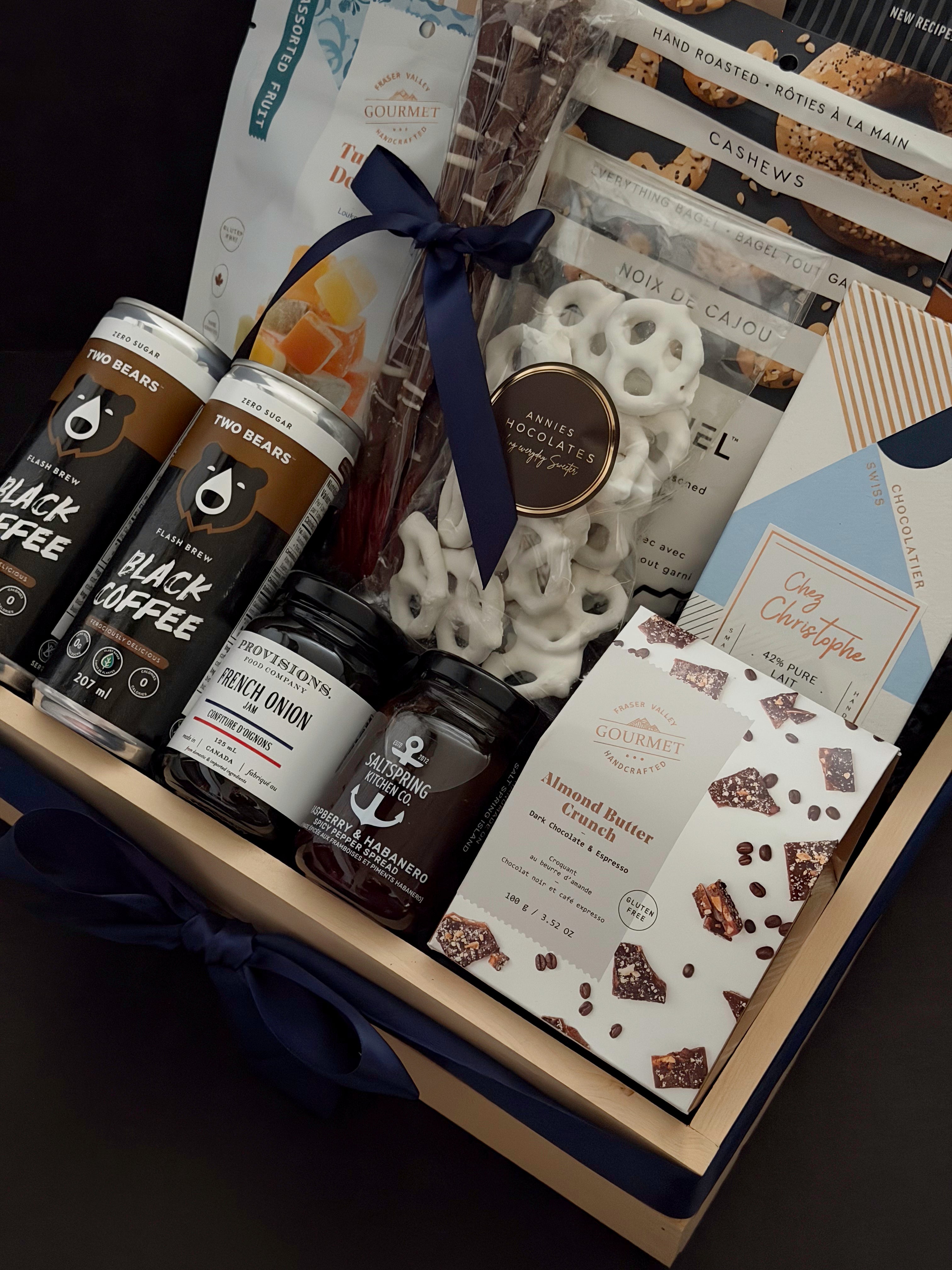 Canadian Gourmet gift box by Muse Baskets and Co., close-up of Two Bears flash brew coffee, Provisions French onion jam, Saltspring Kitchen Co. raspberry habanero spread, Fraser Valley Gourmet almond butter crunch, and white chocolate pretzels in a navy-ribboned wood crate, a luxury Canadian gift for closings and corporate occasions.