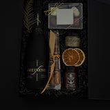 Flat lay of Obsidian Celebration Box contents including sparkling wine, chocolates, pretzels, honey, and seasonal citrus accents.