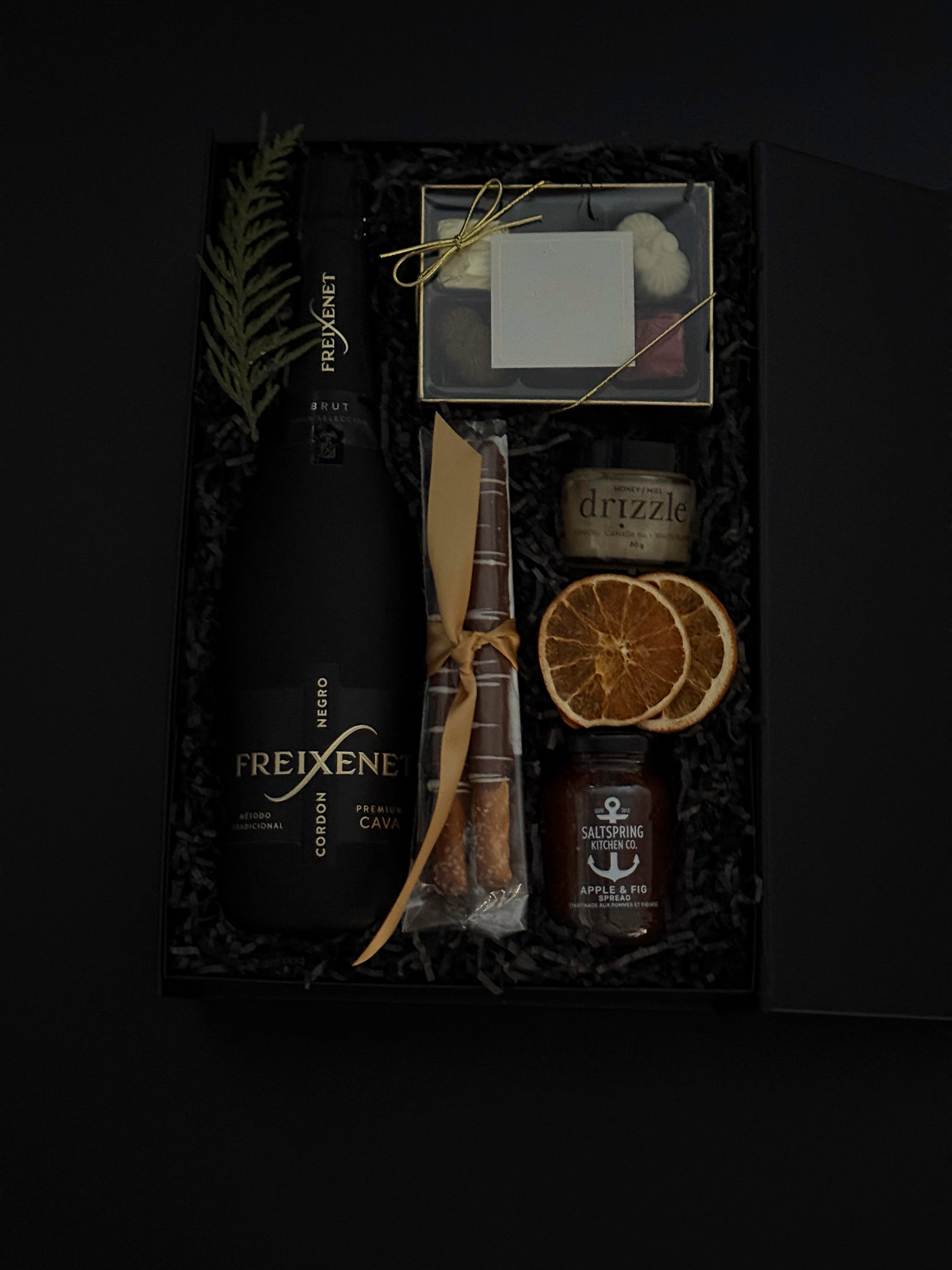 Flat lay of Obsidian Celebration Box contents including sparkling wine, chocolates, pretzels, honey, and seasonal citrus accents.