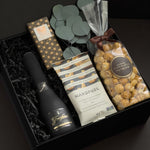 Beaumont gift box by Muse Baskets and Co. shown at an angle, featuring Freixenet Brut Cava, a Laura Slack artisan chocolate bar in gold patterned wrap, Handfuel everything bagel cashews, and Annie's caramel popcorn with eucalyptus on black fill in a black box, a refined corporate gift, client gift, and congratulations gift.