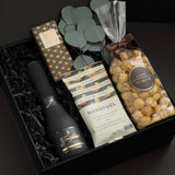 Beaumont gift box by Muse Baskets and Co. shown at an angle, featuring Freixenet Brut Cava, a Laura Slack artisan chocolate bar in gold patterned wrap, Handfuel everything bagel cashews, and Annie's caramel popcorn with eucalyptus on black fill in a black box, a refined corporate gift, client gift, and congratulations gift.