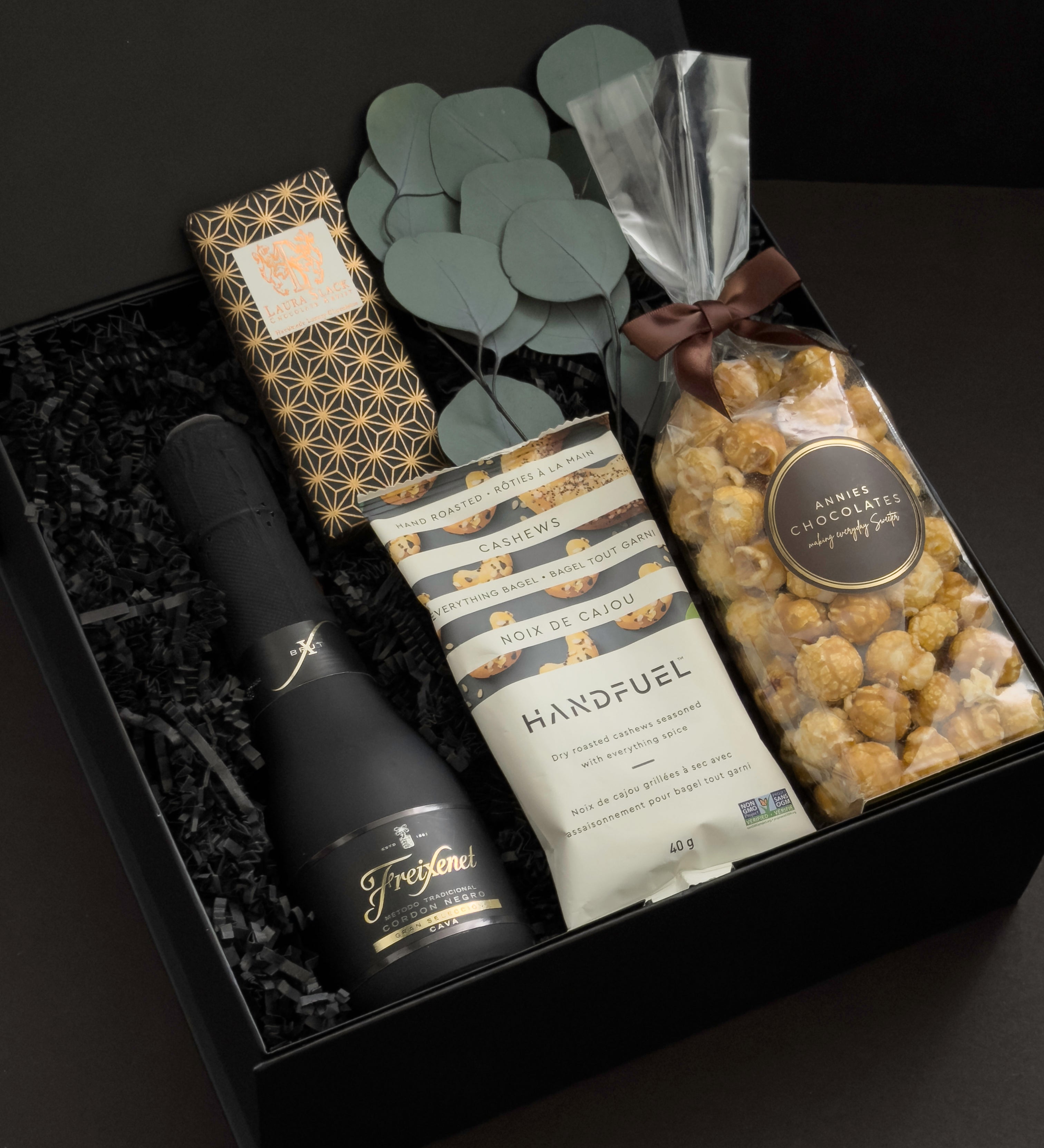 Beaumont gift box by Muse Baskets and Co. shown at an angle, featuring Freixenet Brut Cava, a Laura Slack artisan chocolate bar in gold patterned wrap, Handfuel everything bagel cashews, and Annie's caramel popcorn with eucalyptus on black fill in a black box, a refined corporate gift, client gift, and congratulations gift.
