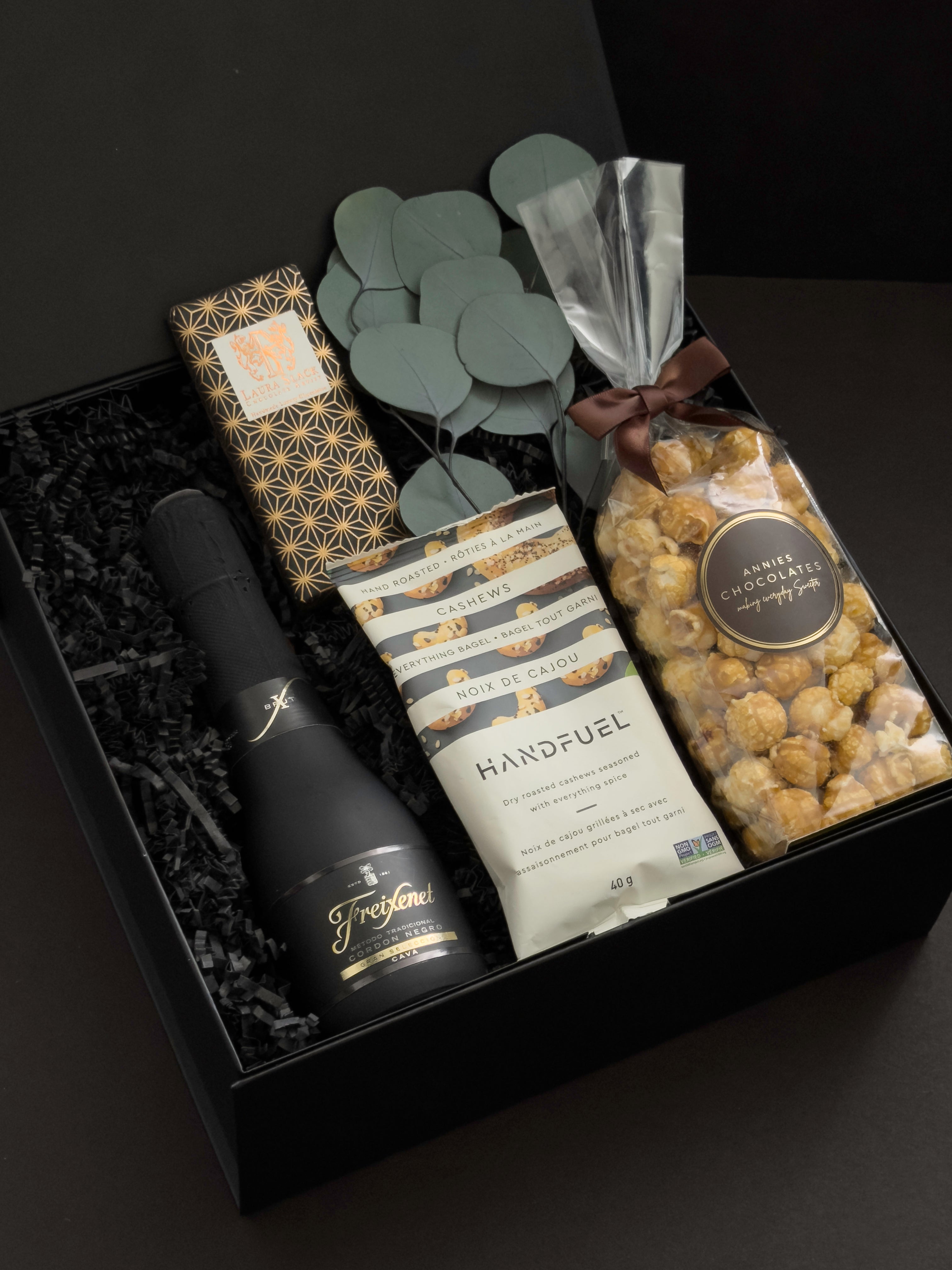 Beaumont gift box by Muse Baskets and Co. shown at an angle, featuring Freixenet Brut Cava, a Laura Slack artisan chocolate bar in gold patterned wrap, Handfuel everything bagel cashews, and Annie's caramel popcorn with eucalyptus on black fill in a black box, a refined corporate gift, client gift, and congratulations gift.