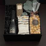 Beaumont gift box by Muse Baskets and Co. photographed straight on, with Freixenet Brut Cava, a Laura Slack chocolate bar, Handfuel everything bagel cashews, and Annie's caramel popcorn tied with a brown ribbon, set with eucalyptus in a black gift box, a curated corporate, referral, and onboarding gift from Canadian makers.