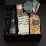 Beaumont gift box by Muse Baskets and Co. photographed straight on, with Freixenet Brut Cava, a Laura Slack chocolate bar, Handfuel everything bagel cashews, and Annie's caramel popcorn tied with a brown ribbon, set with eucalyptus in a black gift box, a curated corporate, referral, and onboarding gift from Canadian makers.