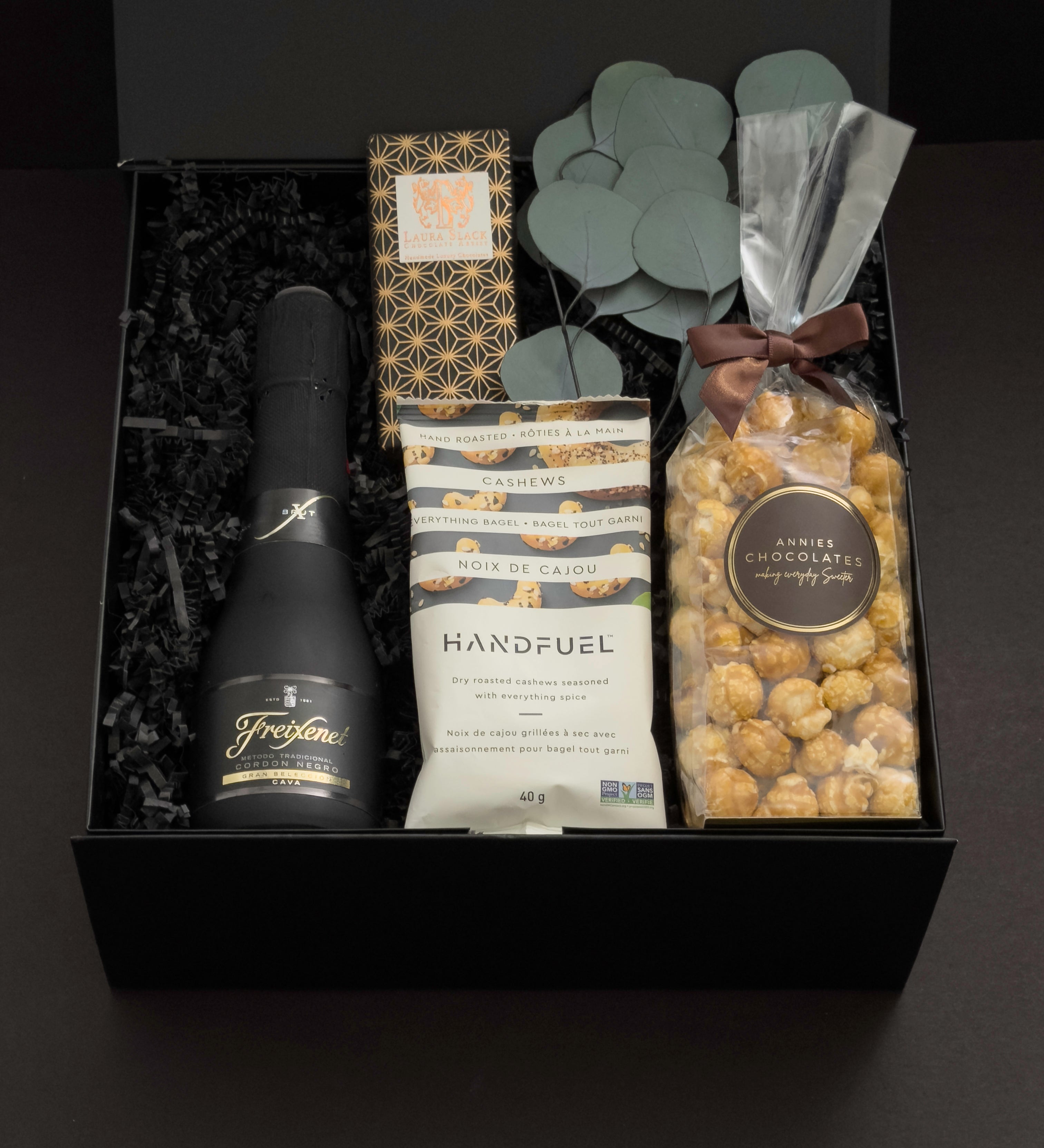 Beaumont gift box by Muse Baskets and Co. photographed straight on, with Freixenet Brut Cava, a Laura Slack chocolate bar, Handfuel everything bagel cashews, and Annie's caramel popcorn tied with a brown ribbon, set with eucalyptus in a black gift box, a curated corporate, referral, and onboarding gift from Canadian makers.