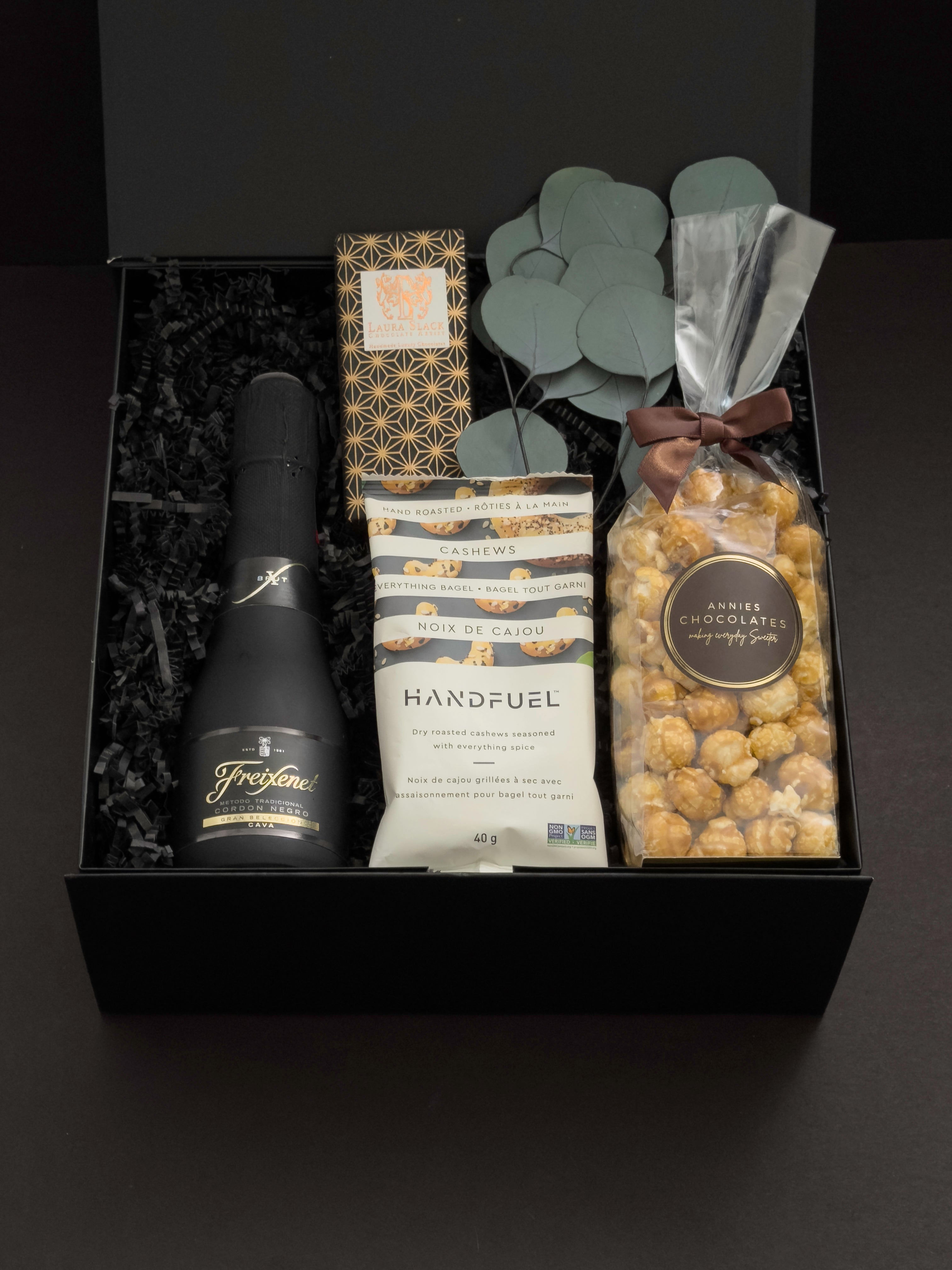 Beaumont gift box by Muse Baskets and Co. photographed straight on, with Freixenet Brut Cava, a Laura Slack chocolate bar, Handfuel everything bagel cashews, and Annie's caramel popcorn tied with a brown ribbon, set with eucalyptus in a black gift box, a curated corporate, referral, and onboarding gift from Canadian makers.
