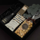 Beaumont gift box by Muse Baskets and Co. viewed close up at an angle, showing Freixenet Brut Cava, a Laura Slack artisan chocolate bar, Handfuel everything bagel cashews, and Annie's caramel popcorn with eucalyptus in a black box, a refined corporate gift, client appreciation gift, and congratulations gift across Canada.