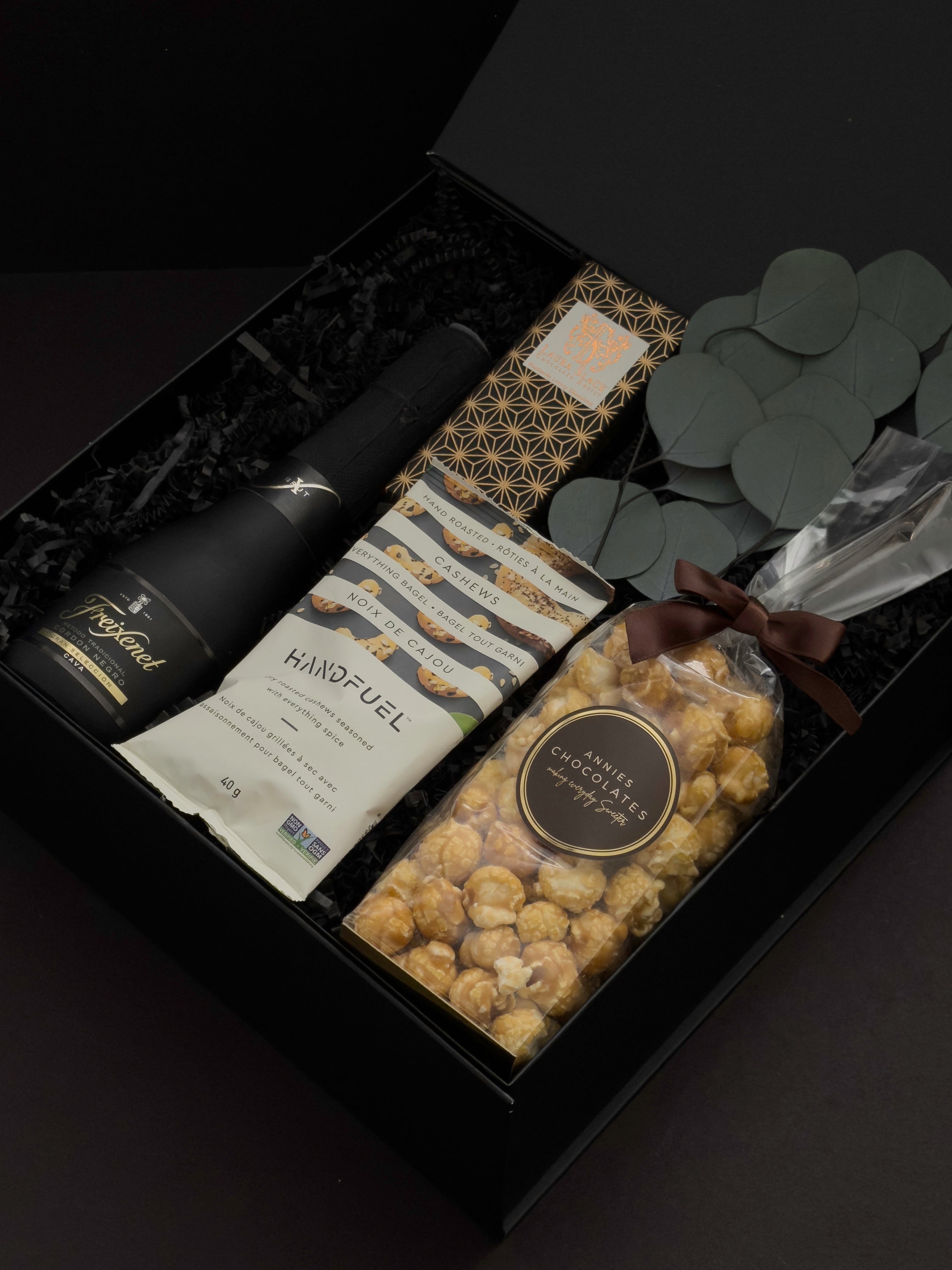 Beaumont gift box by Muse Baskets and Co. viewed close up at an angle, showing Freixenet Brut Cava, a Laura Slack artisan chocolate bar, Handfuel everything bagel cashews, and Annie's caramel popcorn with eucalyptus in a black box, a refined corporate gift, client appreciation gift, and congratulations gift across Canada.