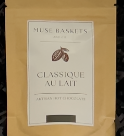 A stand-up pouch of Muse Baskets 'Classique au Lait' artisan hot chocolate, featuring a beige kraft paper exterior and a white label with brown cocoa bean graphics.