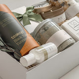 Élysée appreciation gift box by Muse Baskets and Co. photographed from above at an angle, with Teakop Black Yunnan tea, a Muse candle, Sealuxe seaweed body scrub, a So Luxury honey oat cleansing bar and wooden scoop, lemon lavender bath tea, and hand lotion in a white box with eucalyptus and olive green ribbon, a thoughtful corporate and client appreciation gift.