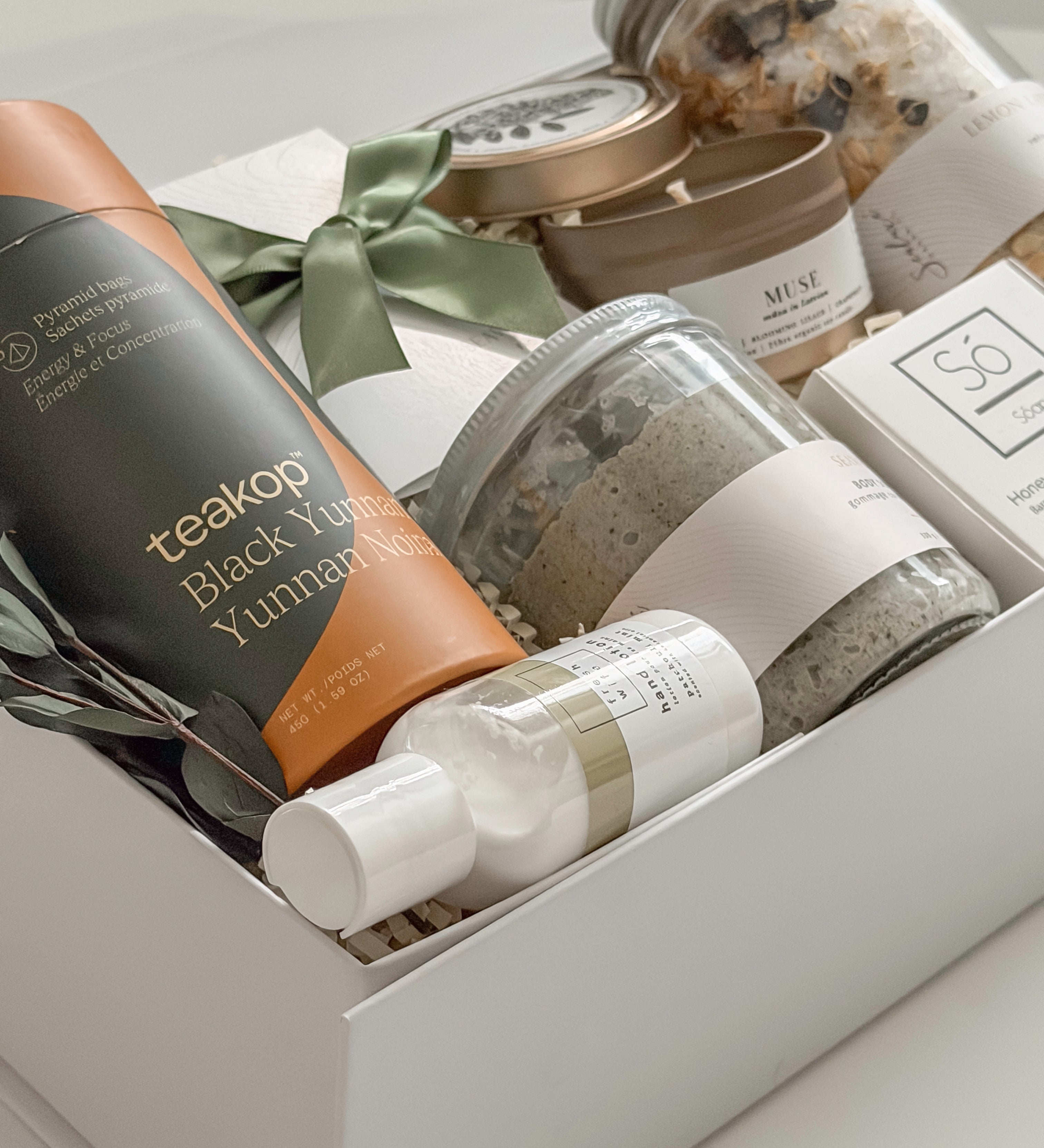 Élysée appreciation gift box by Muse Baskets and Co. photographed from above at an angle, with Teakop Black Yunnan tea, a Muse candle, Sealuxe seaweed body scrub, a So Luxury honey oat cleansing bar and wooden scoop, lemon lavender bath tea, and hand lotion in a white box with eucalyptus and olive green ribbon, a thoughtful corporate and client appreciation gift.