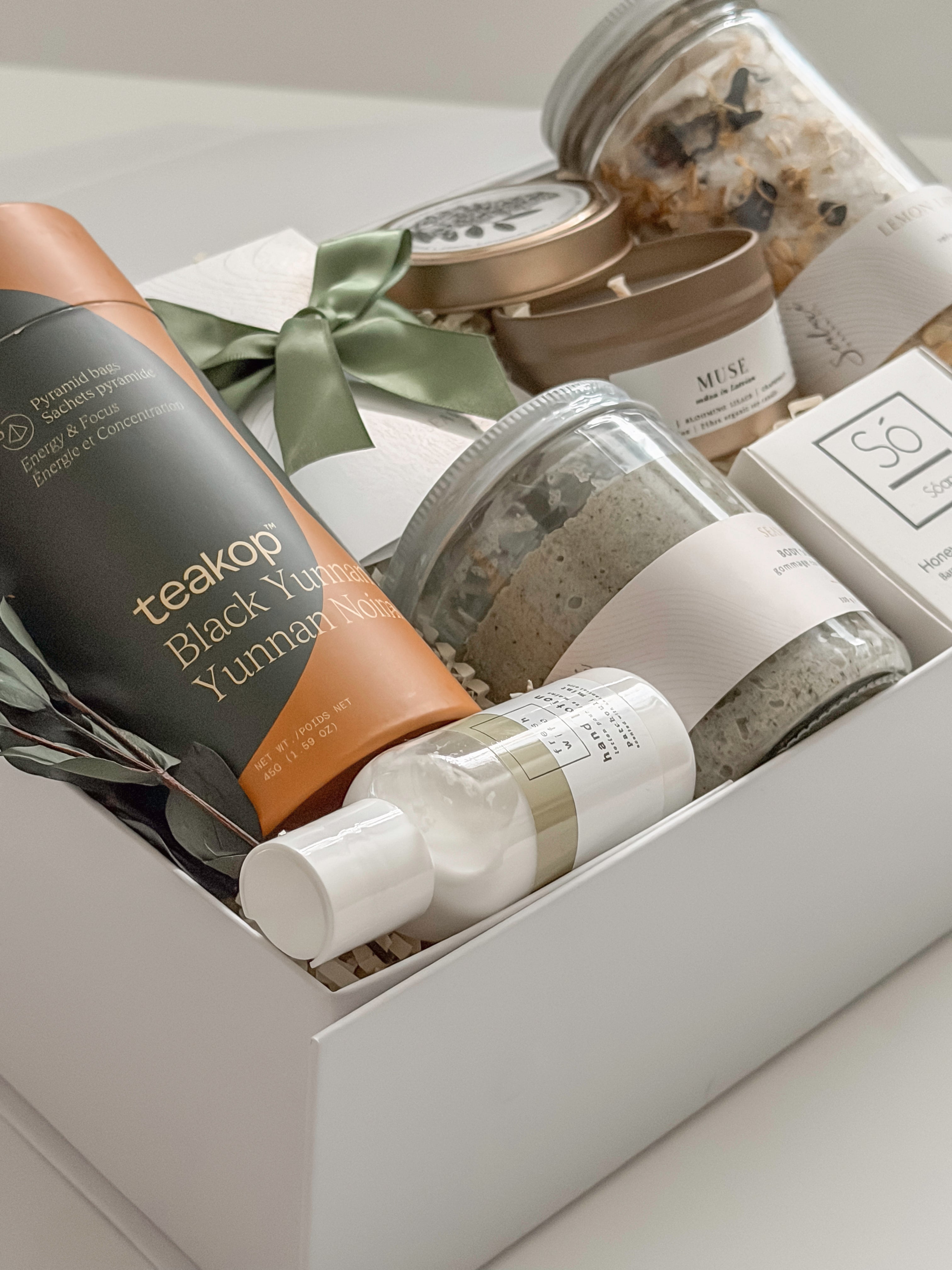 Élysée appreciation gift box by Muse Baskets and Co. photographed from above at an angle, with Teakop Black Yunnan tea, a Muse candle, Sealuxe seaweed body scrub, a So Luxury honey oat cleansing bar and wooden scoop, lemon lavender bath tea, and hand lotion in a white box with eucalyptus and olive green ribbon, a thoughtful corporate and client appreciation gift.