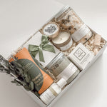 Élysée appreciation gift box by Muse Baskets and Co. viewed close up, with Teakop Black Yunnan tea, Sealuxe seaweed body scrub, a So Luxury honey oat cleansing bar and wooden scoop, a Muse candle, lemon lavender bath tea, and hand lotion arranged in a white box with eucalyptus and an olive green ribbon, a curated corporate and Administrative Professionals Day gift from Canadian makers.