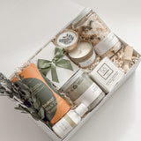 Élysée appreciation gift box by Muse Baskets and Co. viewed close up, with Teakop Black Yunnan tea, Sealuxe seaweed body scrub, a So Luxury honey oat cleansing bar and wooden scoop, a Muse candle, lemon lavender bath tea, and hand lotion arranged in a white box with eucalyptus and an olive green ribbon, a curated corporate and Administrative Professionals Day gift from Canadian makers.