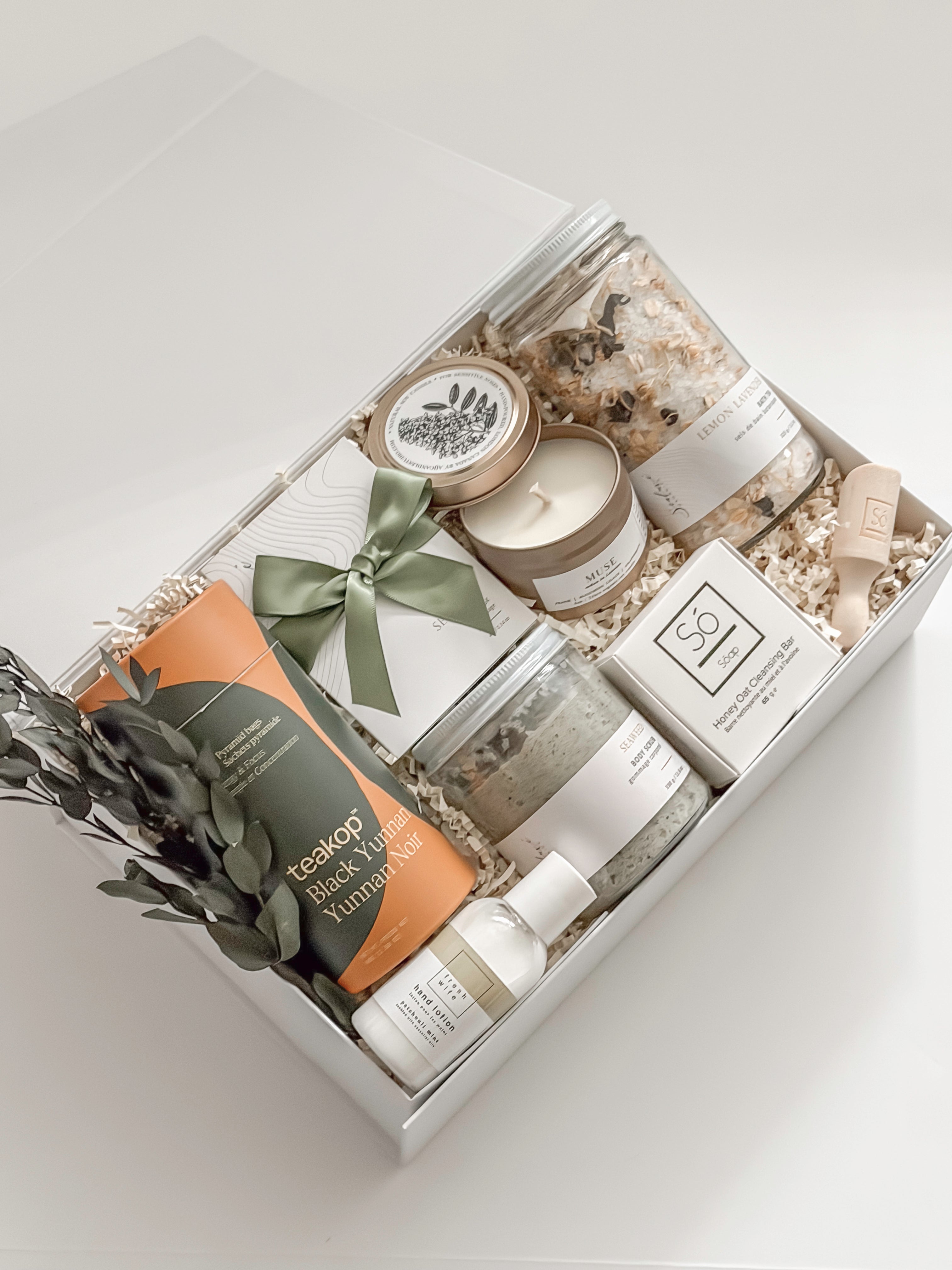 Élysée appreciation gift box by Muse Baskets and Co. viewed close up, with Teakop Black Yunnan tea, Sealuxe seaweed body scrub, a So Luxury honey oat cleansing bar and wooden scoop, a Muse candle, lemon lavender bath tea, and hand lotion arranged in a white box with eucalyptus and an olive green ribbon, a curated corporate and Administrative Professionals Day gift from Canadian makers.