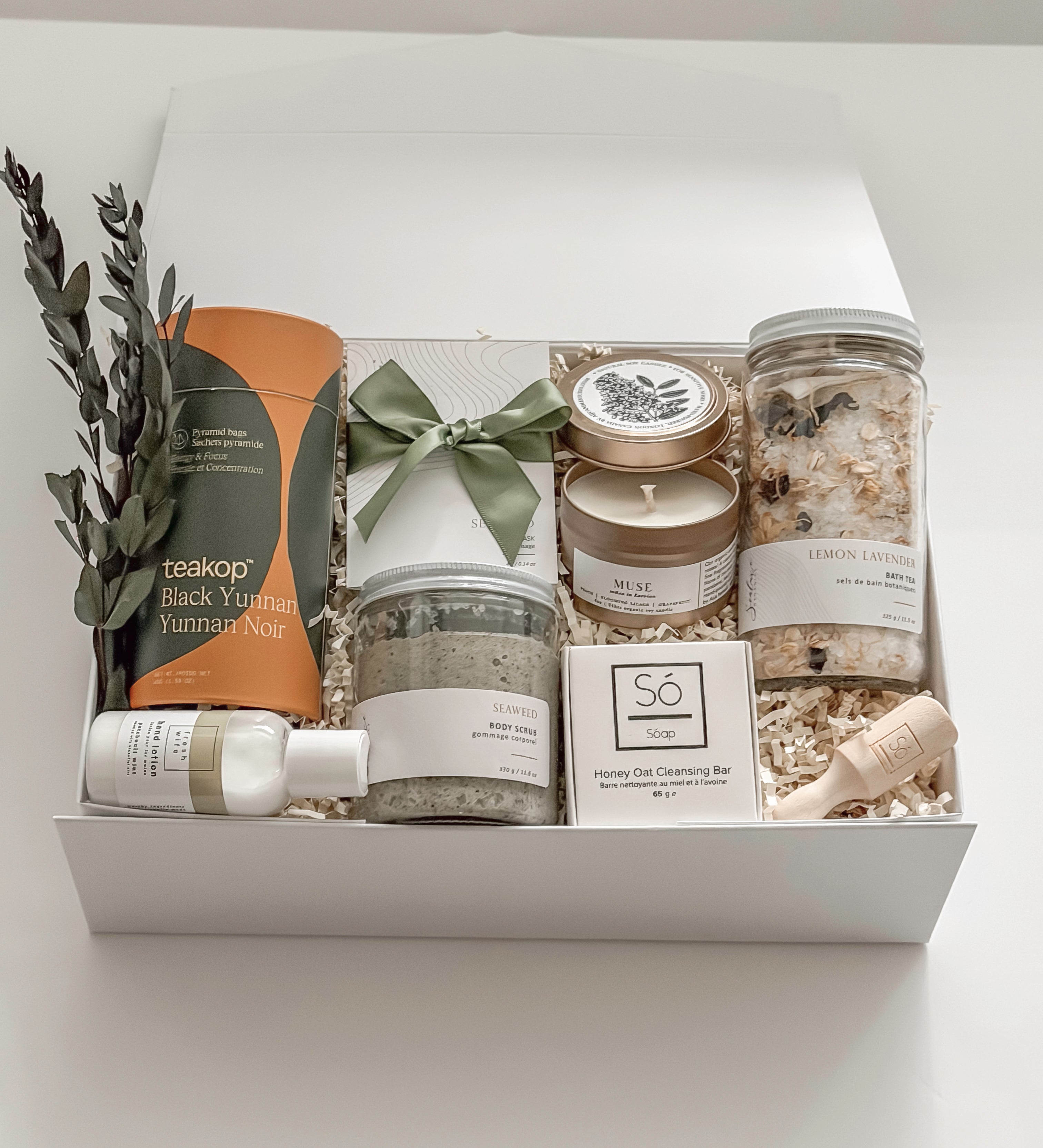 Élysée spa gift box by Muse Baskets and Co. shown close up at an angle, featuring Teakop Black Yunnan tea, a Muse candle by Aija, Sealuxe seaweed body scrub and face mask, Fresh Wife patchouli mint hand lotion, and lemon lavender bath tea with eucalyptus and an olive green ribbon, a refined thank you and employee appreciation gift from Canadian makers.