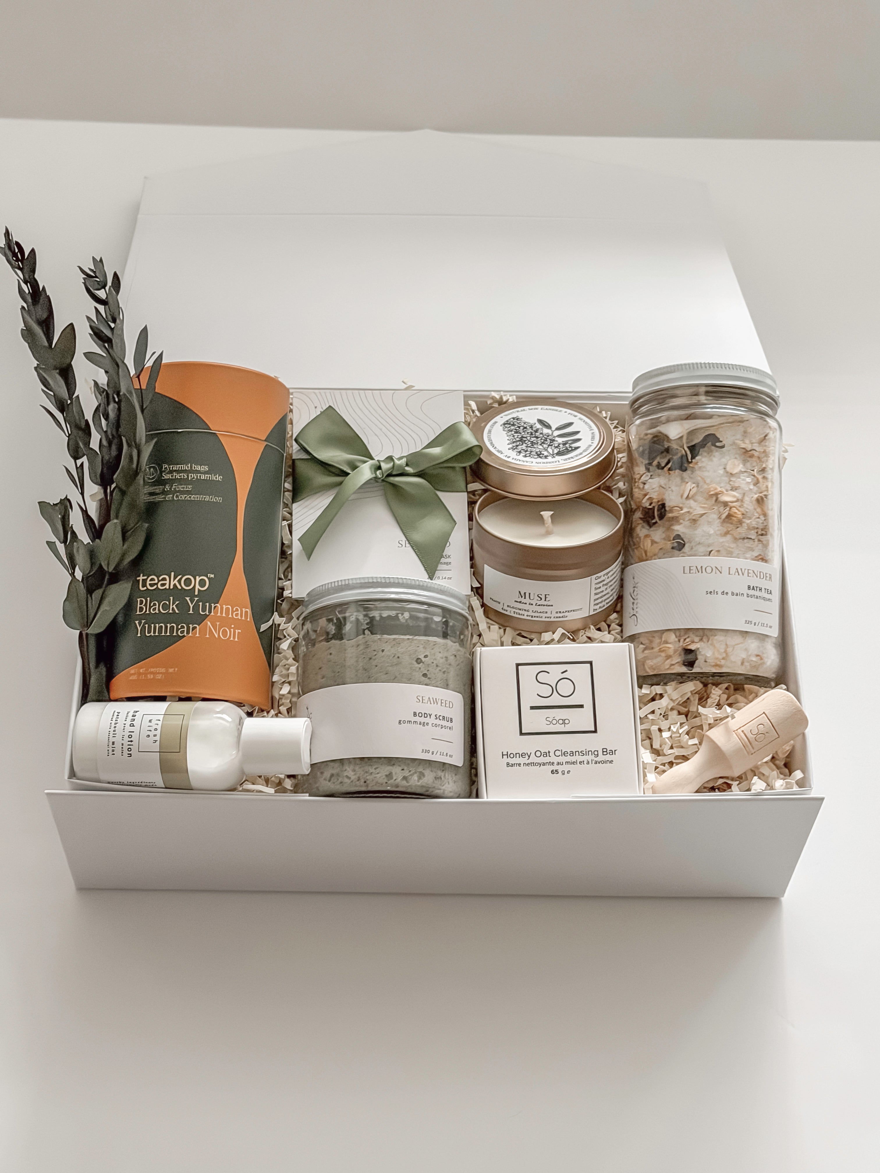 Élysée spa gift box by Muse Baskets and Co. shown close up at an angle, featuring Teakop Black Yunnan tea, a Muse candle by Aija, Sealuxe seaweed body scrub and face mask, Fresh Wife patchouli mint hand lotion, and lemon lavender bath tea with eucalyptus and an olive green ribbon, a refined thank you and employee appreciation gift from Canadian makers.