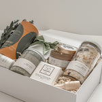 Élysée spa gift box by Muse Baskets and Co. shown from above, featuring Teakop Black Yunnan tea, a Muse candle by Aija, Sealuxe seaweed body scrub and face mask and lemon lavender bath tea, a So Luxury honey oat cleansing bar and wooden scoop, and patchouli mint hand lotion in a white box with eucalyptus and olive green ribbon, a refined thank you, employee appreciation, and client appreciation gift.