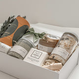 Élysée spa gift box by Muse Baskets and Co. shown from above, featuring Teakop Black Yunnan tea, a Muse candle by Aija, Sealuxe seaweed body scrub and face mask and lemon lavender bath tea, a So Luxury honey oat cleansing bar and wooden scoop, and patchouli mint hand lotion in a white box with eucalyptus and olive green ribbon, a refined thank you, employee appreciation, and client appreciation gift.